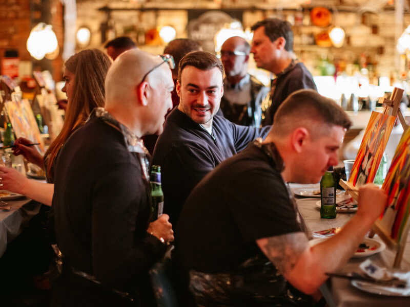 Two men are chatting whilst surrounded by other painters at a drink and paint event.